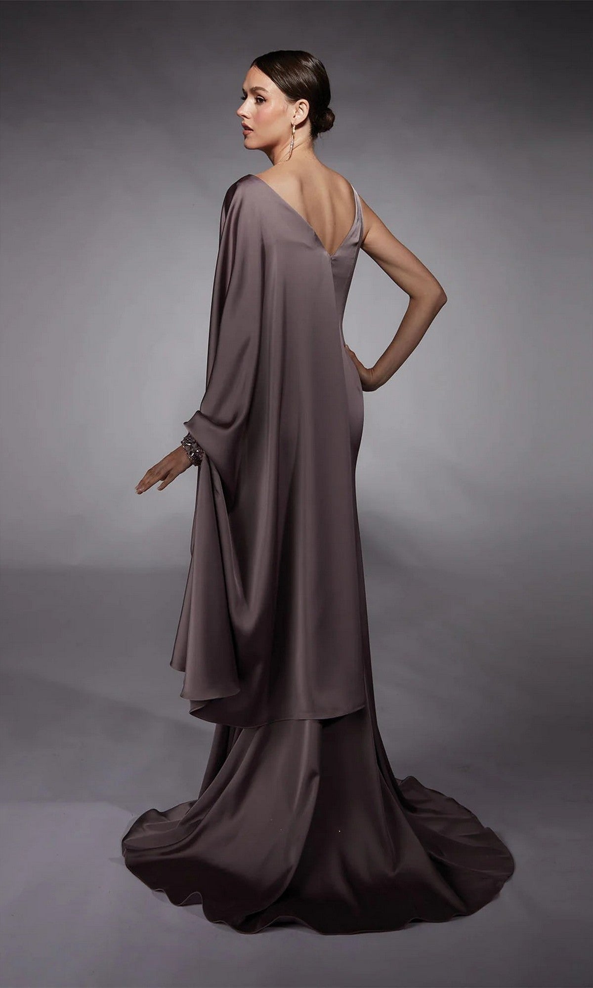 A woman poses with one hand on her hip, looking over her shoulder in the Alyce 27722 long formal dress—a floor-length mauve gown with a draped cape—set against a gray background.